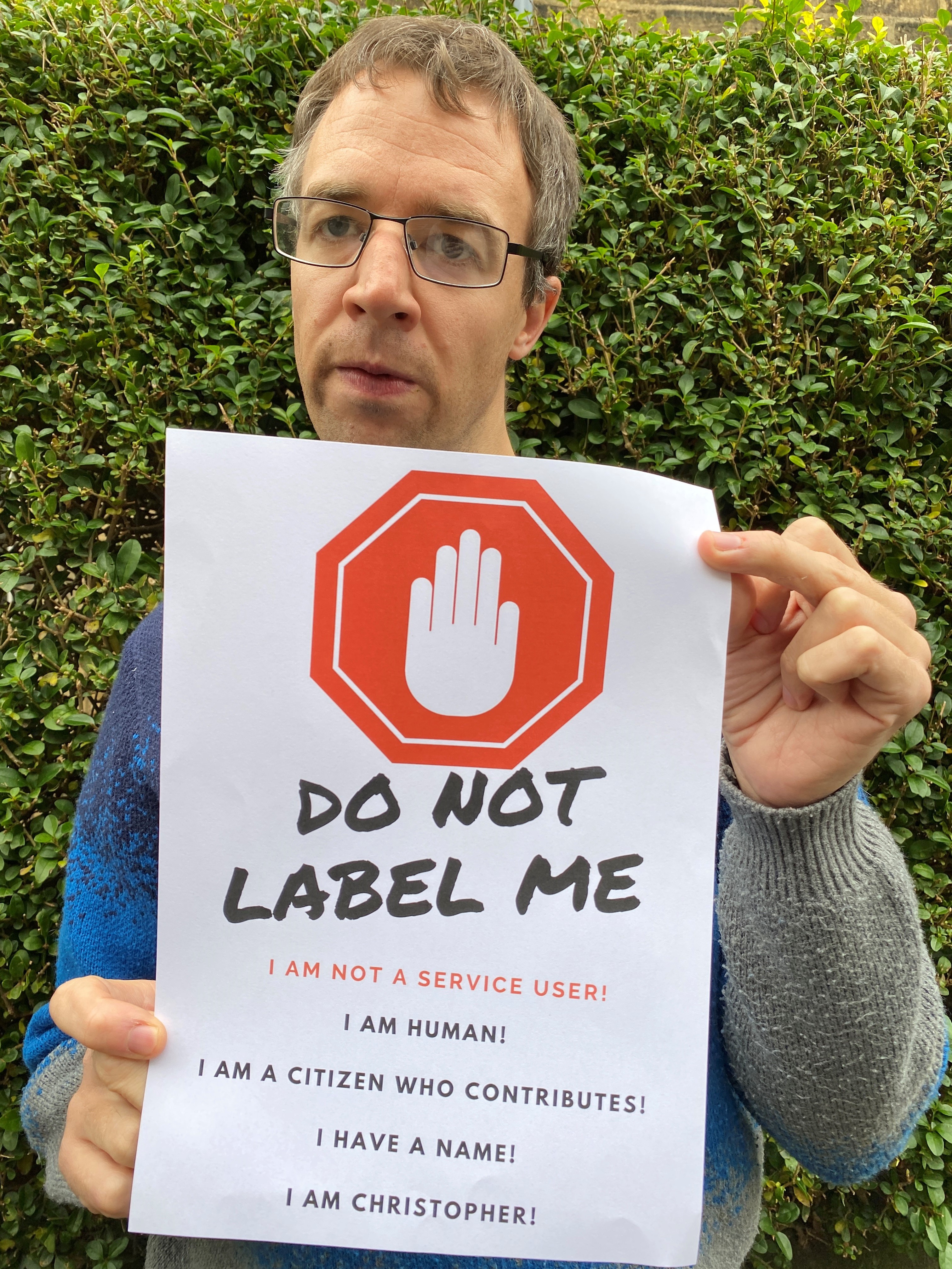 A photograph of Christopher holding a poster. The poster says 'Do not label me. I am not a service user! I am human! I am a citizen who contributes! I have a name! I am Christopher!
