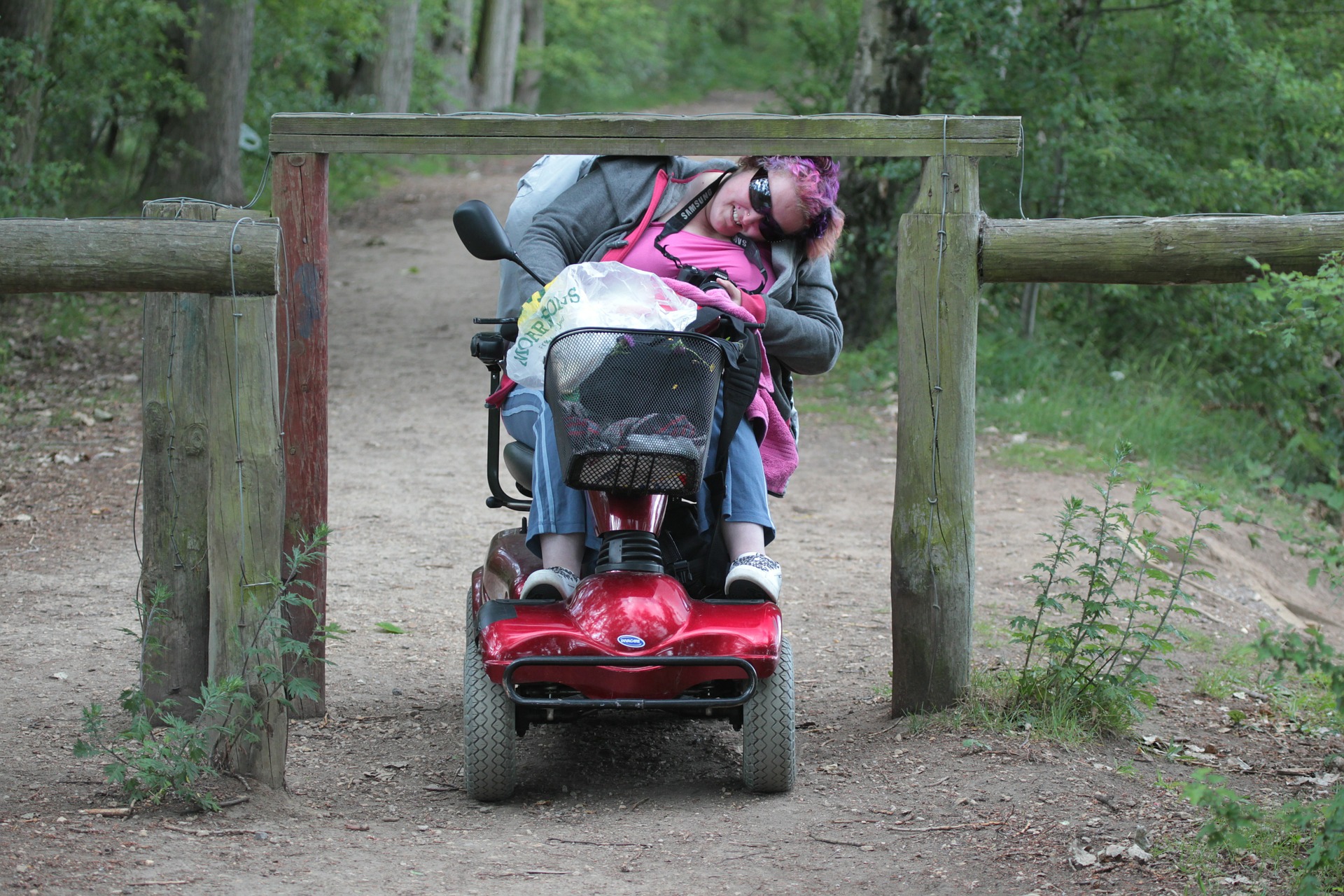 Photo of a person using a mobility scooter on a country path. The person is having to bend over because there is a wooden bar across the path.